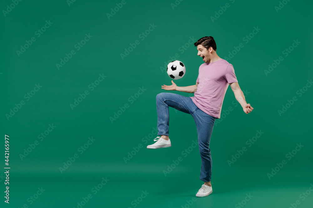 Full size body length young fun man fan wears basic pink t-shirt cheer up support football sport team juggling soccer ball on knee watch tv live stream isolated on dark green color background studio. Full size body length young fun man fan wears basic pink t-shirt cheer up support football sport team juggling soccer ball on knee watch tv live stream isolated on dark green color background studio.