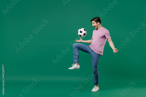 Full size body length young fun man fan wears basic pink t-shirt cheer up support football sport team juggling soccer ball on knee watch tv live stream isolated on dark green color background studio.
