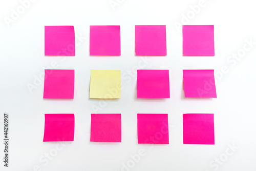 Rows of many colorful bright empty pink sticky paper notes and one stand of a kind stand yellow sticker isolated on white wall textured background. Attached blank memo mockup to do notepad