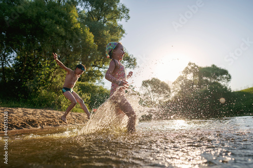 Happy children run from the shore into the water. Summer children s vacation on shore of a lake or river. boy and girl jump into water  swim and splash around at sunset. Active holidays. Dynamic image