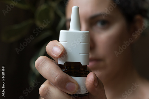 Φωτογραφία Woman holds out allergy nasal spray towards camera; background has green housepl