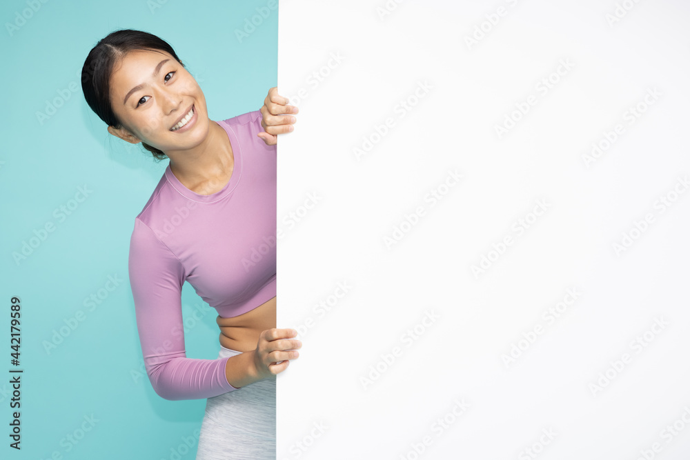 Happy Asian woman in yoga suit standing behind blank white billboard Happy Asian woman in yoga suit standing behind blank white billboard