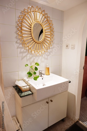 Simple powder room with vanity sink. There is rattan bamboo mirror on the wall, white countertop and cabinet, there is green vine plants pot
