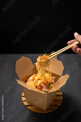 Noodle with pork, meat, vegetable in cardboard box for takeaway with chopstick in dark or black background, asian traditional chinese cuisine food for delivery concept with copy space