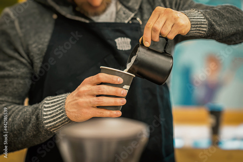 Unrecognizable Barista wearing black apron making cappuccino, pouring milk in paper cup in coffee bar