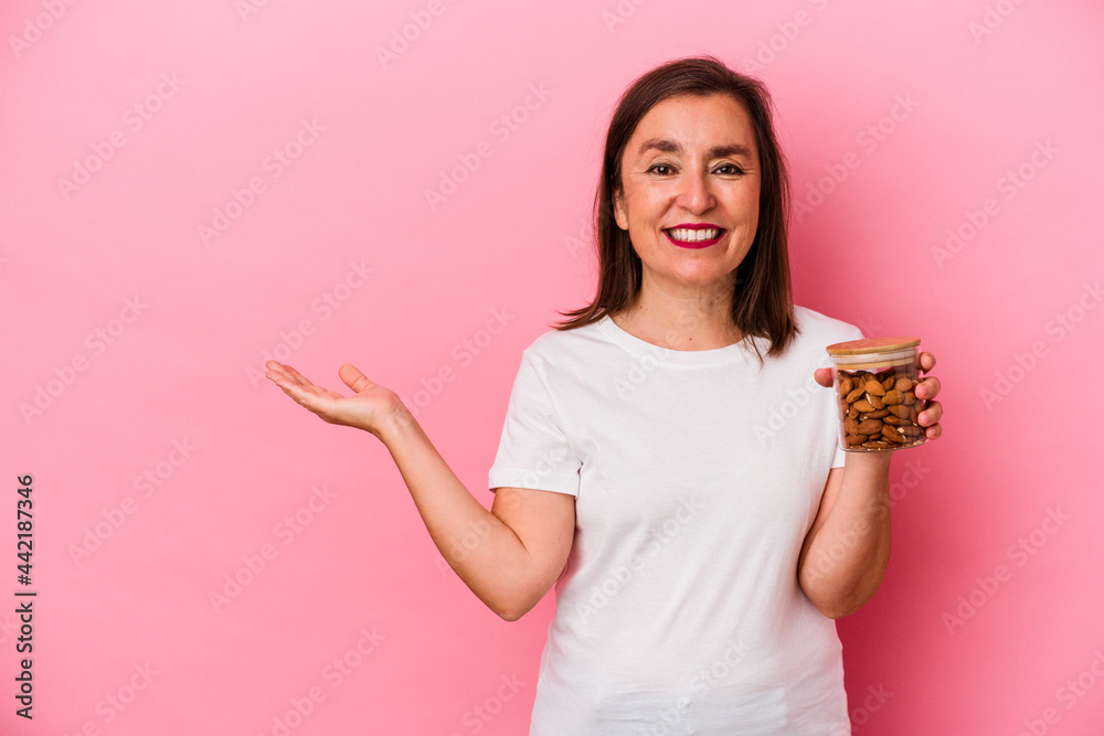 Middle age caucasian woman holding an almond jar isolated on pink background showing a copy space on a palm and holding another hand on waist. Middle age caucasian woman holding an almond jar isolated on pink background showing a copy space on a palm and holding another hand on waist.