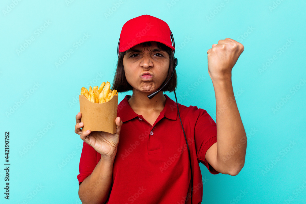 Young mixed race woman fast food restaurant worker holding fries isolated on blue background showing fist to camera, aggressive facial expression. Young mixed race woman fast food restaurant worker holding fries isolated on blue background showing fist to camera, aggressive facial expression.