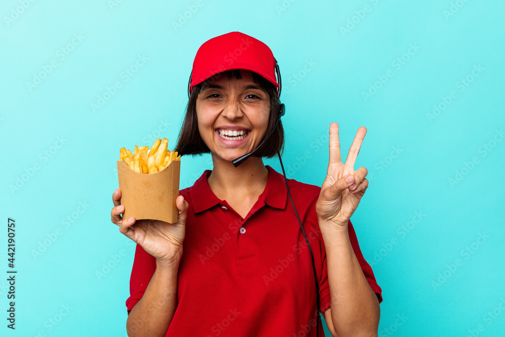 Young mixed race woman fast food restaurant worker holding fries isolated on blue background joyful and carefree showing a peace symbol with fingers. Young mixed race woman fast food restaurant worker holding fries isolated on blue background joyful and carefree showing a peace symbol with fingers.
