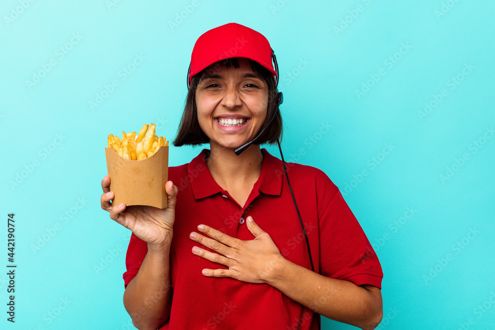 Young mixed race woman fast food restaurant worker holding fries isolated on blue background laughs out loudly keeping hand on chest. Young mixed race woman fast food restaurant worker holding fries isolated on blue background laughs out loudly keeping hand on chest.