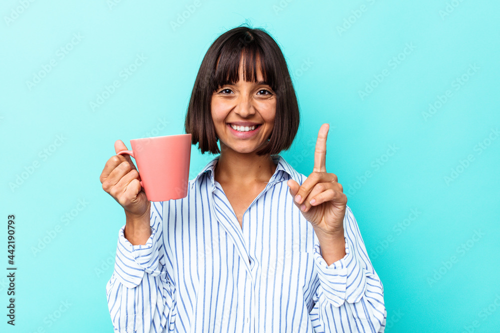 Young mixed race woman holding a pink mug isolated on blue background showing number one with finger. Young mixed race woman holding a pink mug isolated on blue background showing number one with finger.