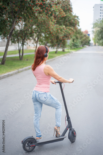A red-haired young woman in ripped jeans and high-heeled sandals rides an electric scooter on the road and listens to music on her bluetooth wireless headphones. Convenient electric transport.
