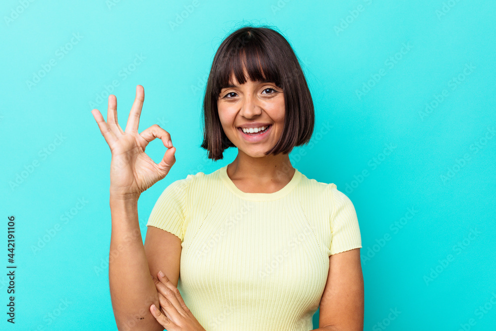 Young mixed race woman isolated on blue background winks an eye and holds an okay gesture with hand. Young mixed race woman isolated on blue background winks an eye and holds an okay gesture with hand.
