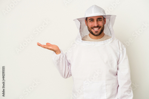 Young apiculture caucasian man isolated on white background showing a copy space on a palm and holding another hand on waist.