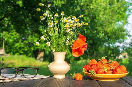 A plate with strawberries and a bouquet of summer flowers and a newspaper with reading glasses on a wooden table in the garden. Summer still life