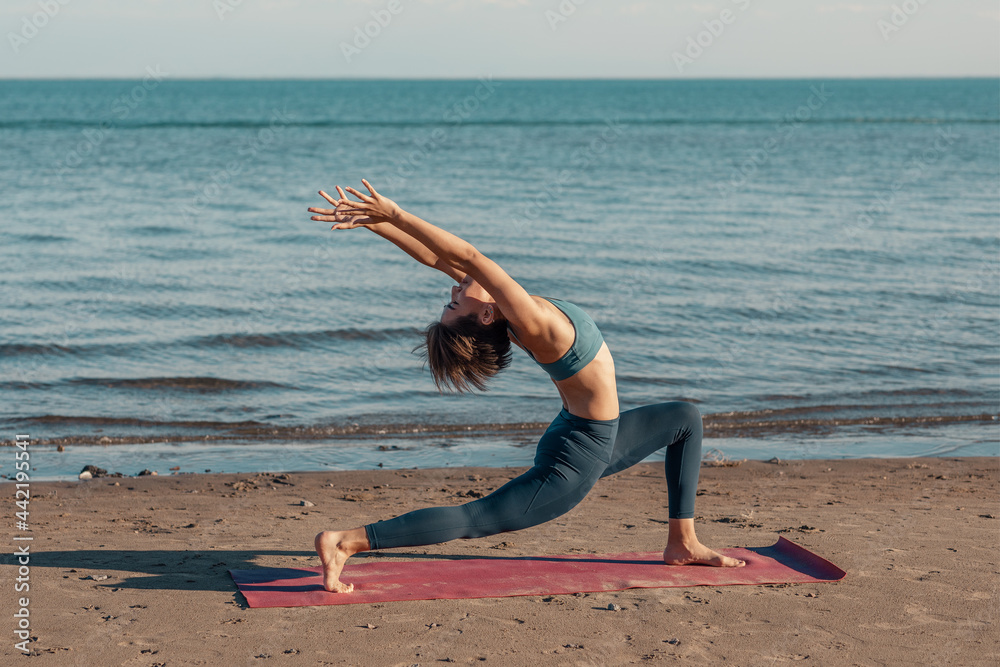 a young attractive woman, of Asian appearance, practicing yoga, performs a stretching exercise, on the beach. the warrior pose with a backward bend