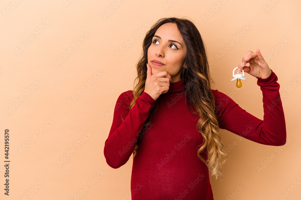 Fototapeta premium Young mexican pregnant woman holding pacifier isolated on beige background looking sideways with doubtful and skeptical expression.
