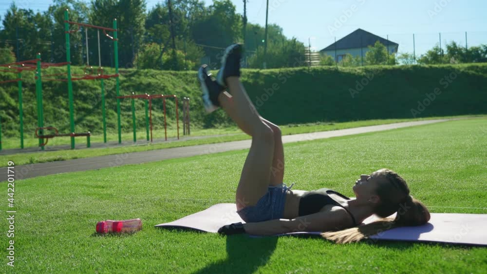 Side motion of sexy muscular young woman training abdominal muscles while lying on mat on green stadium field in sunny summer day. Fitnessgirl raising legs up outdoors. Workout concept. Side motion of sexy muscular young woman training abdominal muscles while lying on mat on green stadium field in sunny summer day. Fitnessgirl raising legs up outdoors. Workout concept.