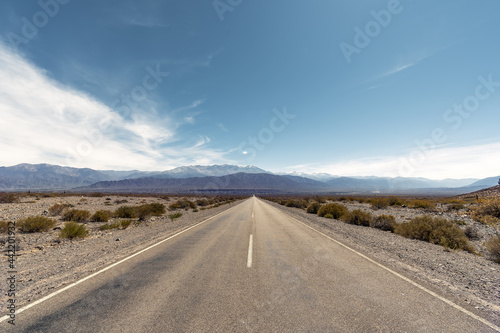 two lane road in a arid landscape in Argentina leading to mountains against blue sky © Marcos Reppetti