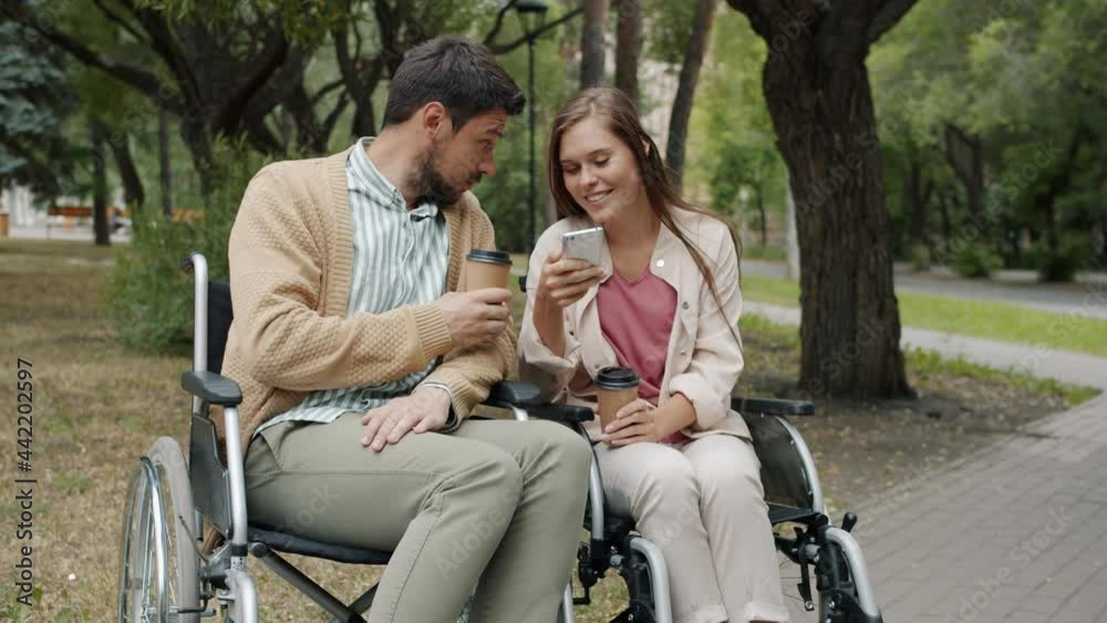 Disabled husband and wife are sitting in wheelchairs talking using smartphone drinking take-out coffee in park on autumn day. Disability and lifestyle concept. Disabled husband and wife are sitting in wheelchairs talking using smartphone drinking take-out coffee in park on autumn day. Disability and lifestyle concept.