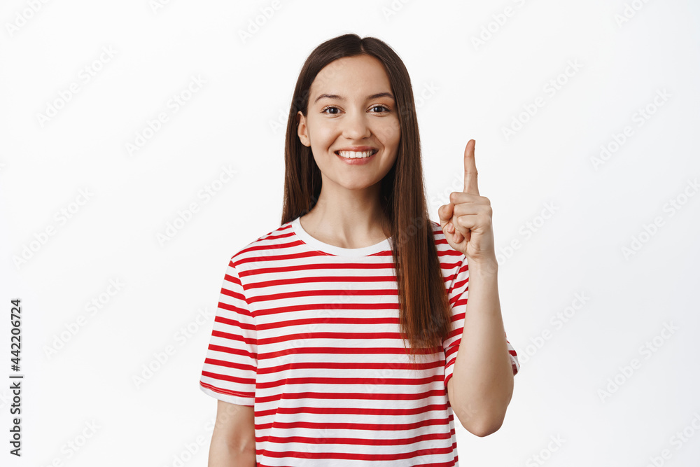 Beautiful smiling girl pointing finger up and looking happy, showing sale, promotional text for your company, standing in tshirt against white background Beautiful smiling girl pointing finger up and looking happy, showing sale, promotional text for your company, standing in tshirt against white background