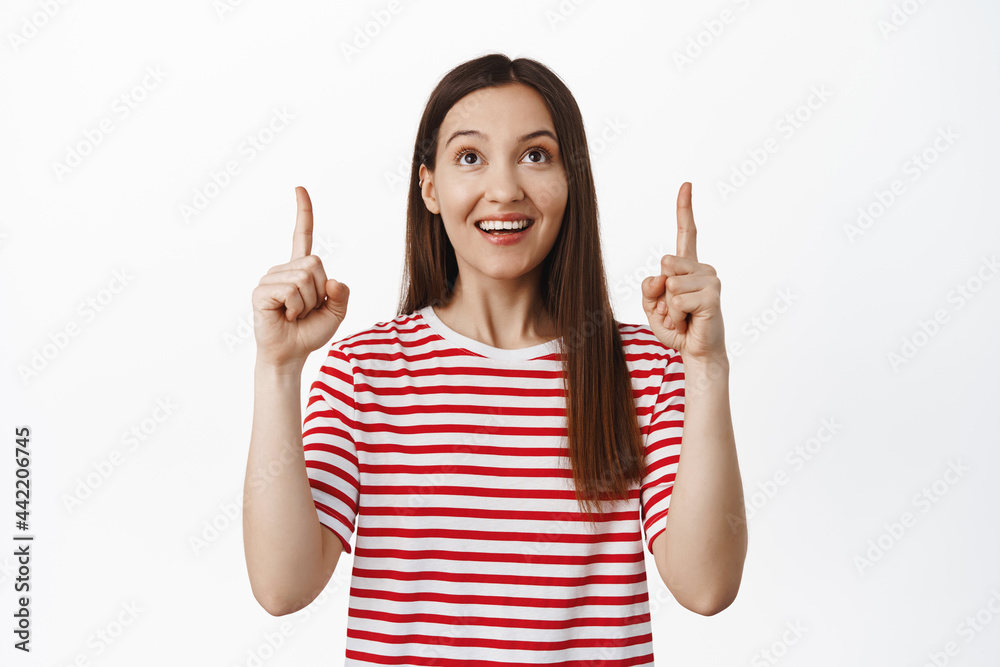 Portrait of beautiful brunette girl pointing, looking up and smiling happy, reading promotional text, showing sale banner, standing in red t-shirt against white background Portrait of beautiful brunette girl pointing, looking up and smiling happy, reading promotional text, showing sale banner, standing in red t-shirt against white background