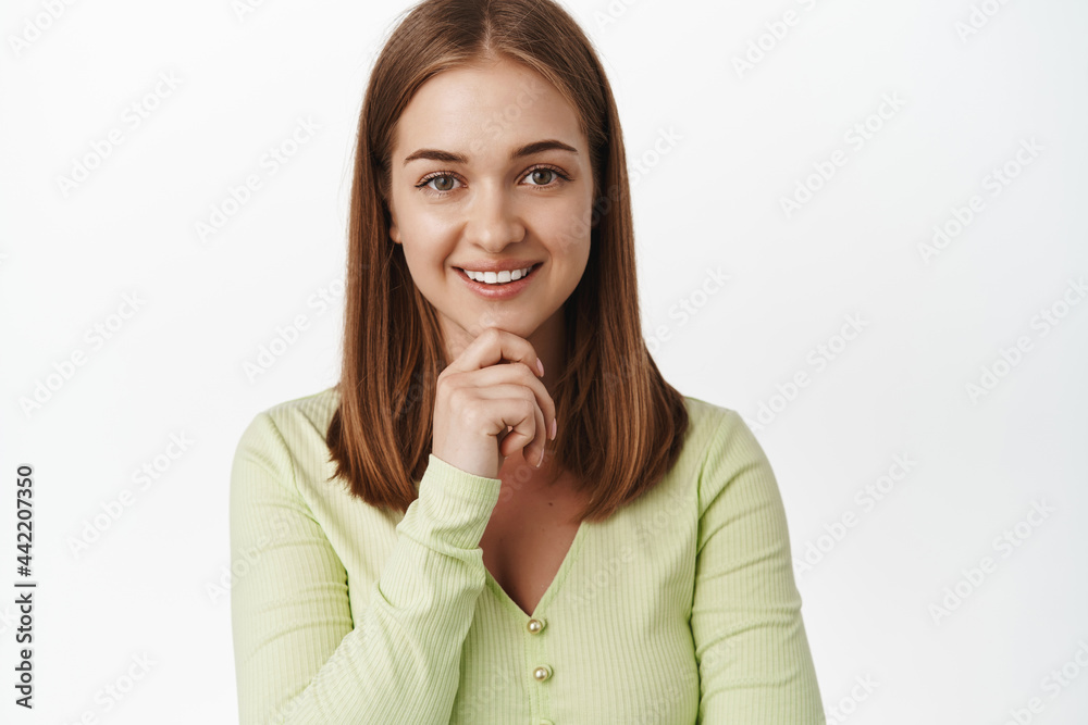 Close up portrait of blond smiling woman, touch chin, look pleased and thoughtful, has an idea, thinking and staring at camera intrigued, standing against white background Close up portrait of blond smiling woman, touch chin, look pleased and thoughtful, has an idea, thinking and staring at camera intrigued, standing against white background