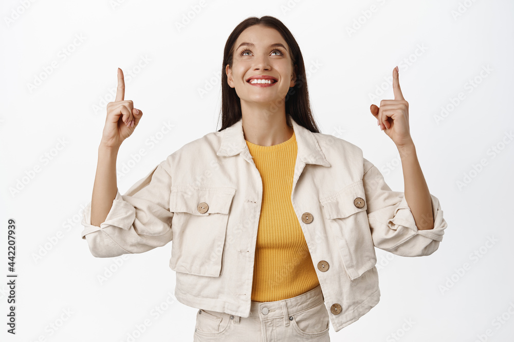 Smiling beautiful woman pointing, looking up with pleased happy face, seeing discount, showing logo banner, place for advertisement, standing in casual clothes against white background Smiling beautiful woman pointing, looking up with pleased happy face, seeing discount, showing logo banner, place for advertisement, standing in casual clothes against white background