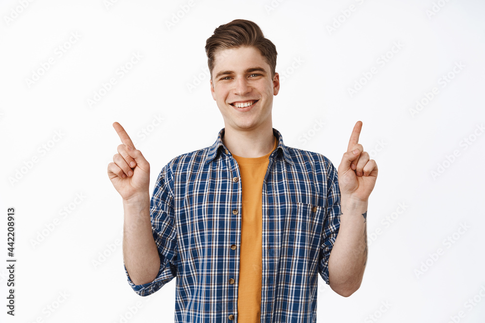 Young attractive smiling guy, college boy pointing fingers aside, showing two product banners, variety of discounts and look happy, standing over white background Young attractive smiling guy, college boy pointing fingers aside, showing two product banners, variety of discounts and look happy, standing over white background