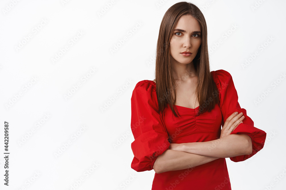 Image of young serious and confident young in red trendy dress, cross arms on chest, looks bossy and self-assured, standing displeased against white background Image of young serious and confident young in red trendy dress, cross arms on chest, looks bossy and self-assured, standing displeased against white background