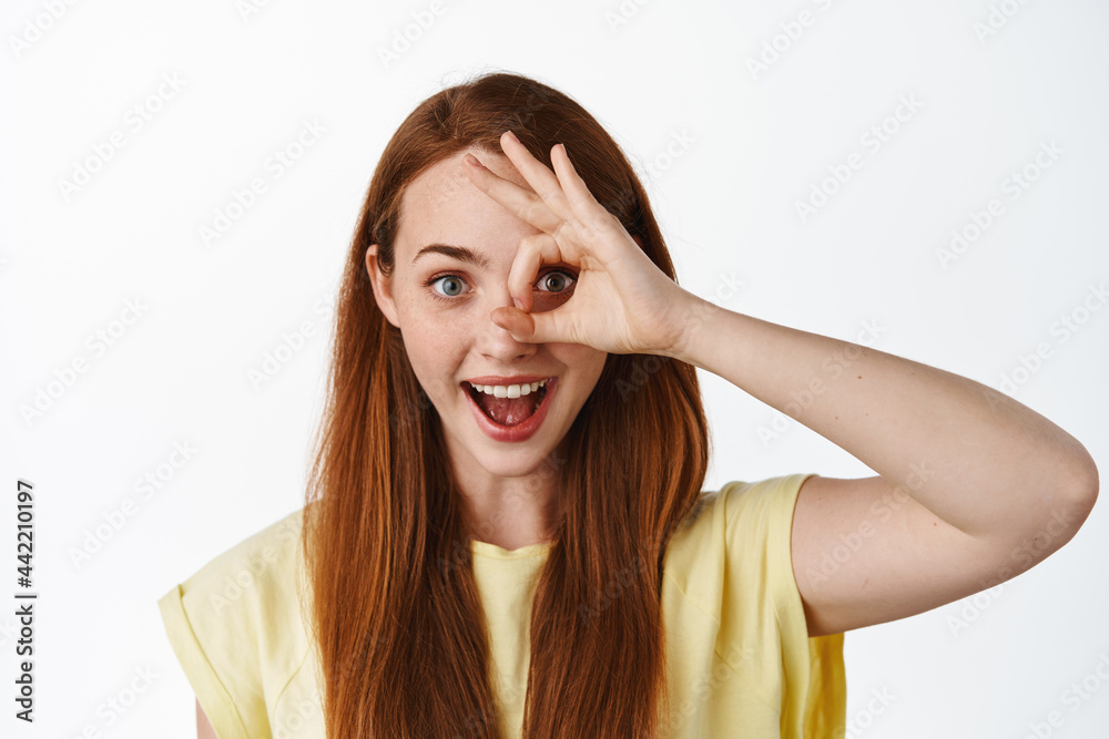 Close up portrait of redhead girl shows zero okay sign, look through OK gesture and smiling amused, checking out something cool, praise you, white background Close up portrait of redhead girl shows zero okay sign, look through OK gesture and smiling amused, checking out something cool, praise you, white background