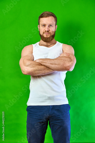 Caucasian man look in camera seriously. Athlete bodybuilder on green background. adult guy with beard in casual clothes white T-shirt and jeans. Looking ahead. Self-confident person.