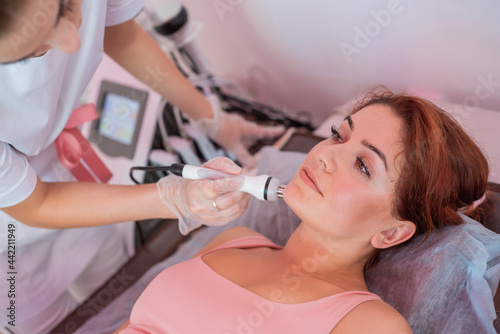 Young redhead woman gets an electric facial massage in the fight against aging. 30 year old woman receives electric ultrasonic facial massage against wrinkles in a beauty salon. Hardware cosmetology.