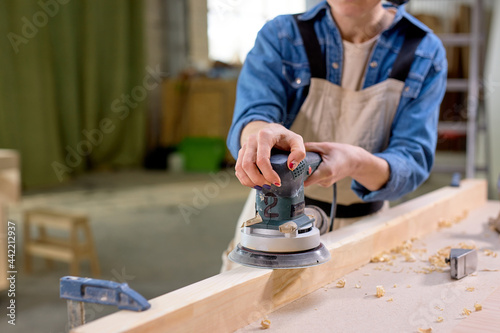 Young cropped female carpenter working with wood using electric sander in workshop. Joinery work on the production and renovation of wooden furniture. Small Business Concept. close-up photo