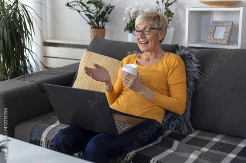 An older woman with short blonde hair sits on sofa in living room and chats with friend on laptop over video chat