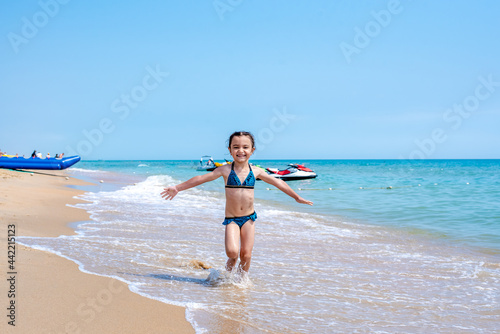 Happy little girl in a bathing suit running with open arms on the golden sand, looking at the camera, spending free time on the beach by the sea. Full length, front view. Childhood concept. Summer vac #442215123