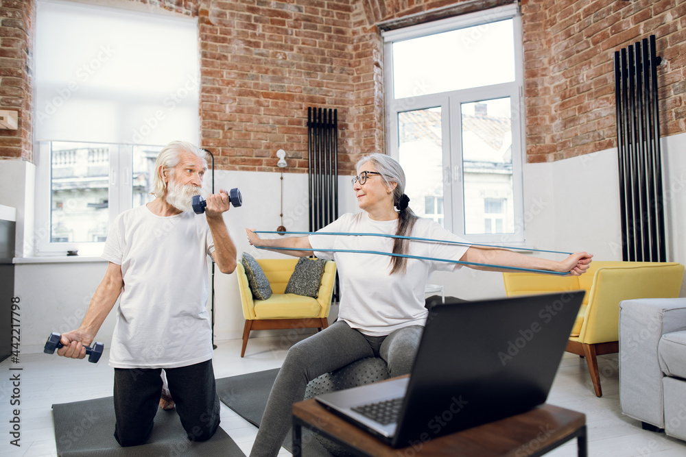 Mature couple in active clothes training with various sport equipments at home. Aged man and woman watching tutorial on modern laptop. Sporty and healthy lifestyles.