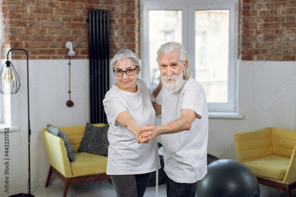 Energetic retired couple in casual clothes moving actively at bright living room. Two happy people dancing together at home. Leisure time concept.