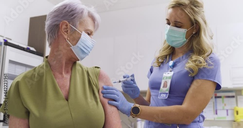 A Mid Shot Of A Female American Medical Nurse Administering A Covid-19, SARS-CoV-2 Vaccine Injection With A Needle and Syringe, To An Elderly Woman. Wearing Safety Gloves And Protective Mask