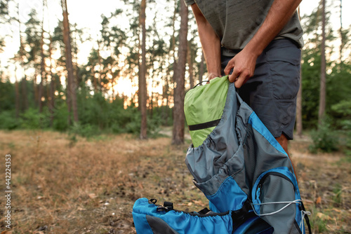 Cropped shot of young male tourist unpacking sleeping bag and other camping equipment, standing in the summer forest