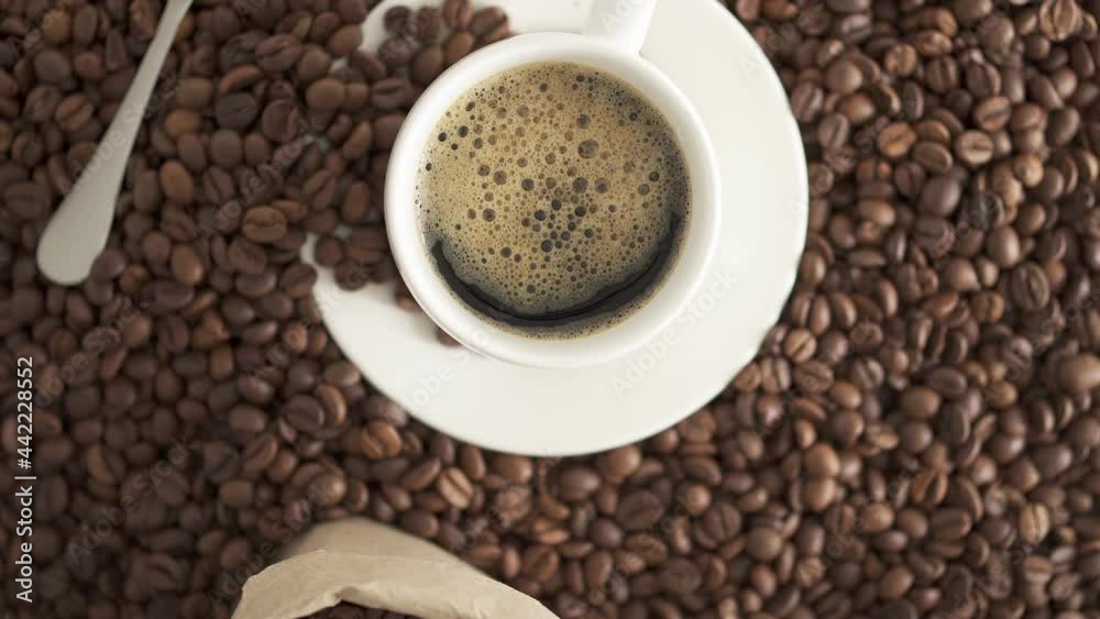 White cup, saucer, spoon on a background of coffee beans. A cup of fresh espresso with froth. Coffee in a craft bag spilled out onto the table. The camera flies around the Arabica beans. Coffee shop White cup, saucer, spoon on a background of coffee beans. A cup of fresh espresso with froth. Coffee in a craft bag spilled out onto the table. The camera flies around the Arabica beans. Coffee shop