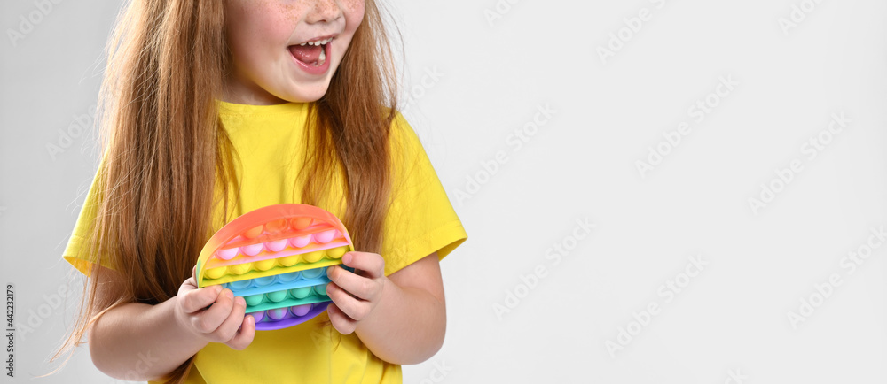 Cropped photo of a red-haired girl with a colorful pop simple silicone toy with dimples shows surprised, looks to the side. Studio face portrait children shot Cropped photo of a red-haired girl with a colorful pop simple silicone toy with dimples shows surprised, looks to the side. Studio face portrait children shot