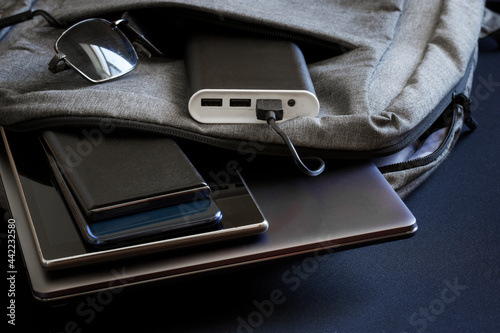 Portable charger - power bank lies in backpack along with sunglasses and gadgets - smartphones, tablet and slim fashionable laptop. Charge your gadgets while on vacation, travel and trip