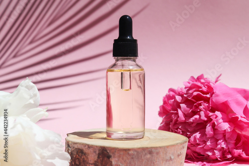 Bottle of cosmetic essential oil with dropper on a wood cut with peony flowers and palm leaf shadow. Beauty concept. Serum skin care product. Selective focus