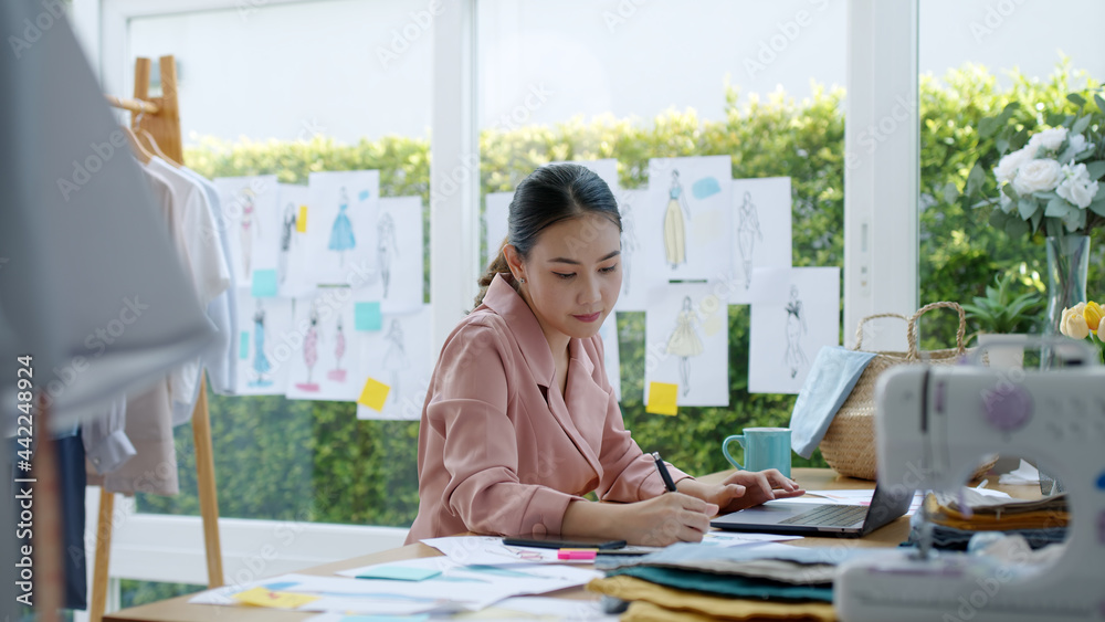 Young happy smile asia success woman busy work on new dress collection on desk laptop tablet at studio workshop workspace home office in sme e-commerce shop sell online store garment textile business.