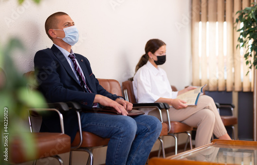 Focused young man in a protective mask, sitting in the lobby of a large company during a pandemic, is waiting for his ..turn to see a specialist