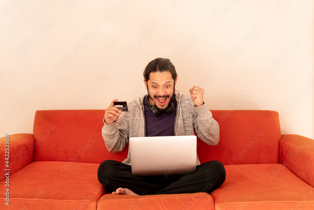 Young man shopping online with credit card at home using computer sitting on an orange couch sofa Young man shopping online with credit card at home using computer sitting on an orange couch sofa