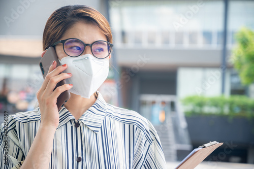 Young Asian woman talking with other by smart phone while wearing a mask for protect herself from virus spreading or bad air pollution. New normal lifestyle of people after covid-19 pandemic.