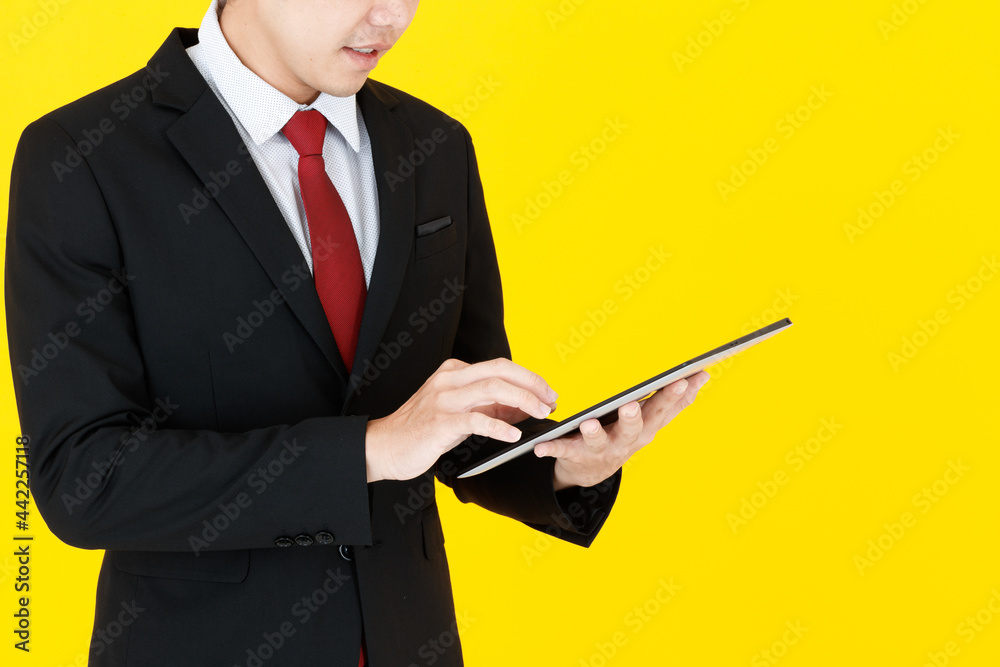 Horizontal portrait shot of young Asian man in a black long sleeve suit holding a tablet while touching the screen for searching for the information isolated with yellow background in the studio Horizontal portrait shot of young Asian man in a black long sleeve suit holding a tablet while touching the screen for searching for the information isolated with yellow background in the studio