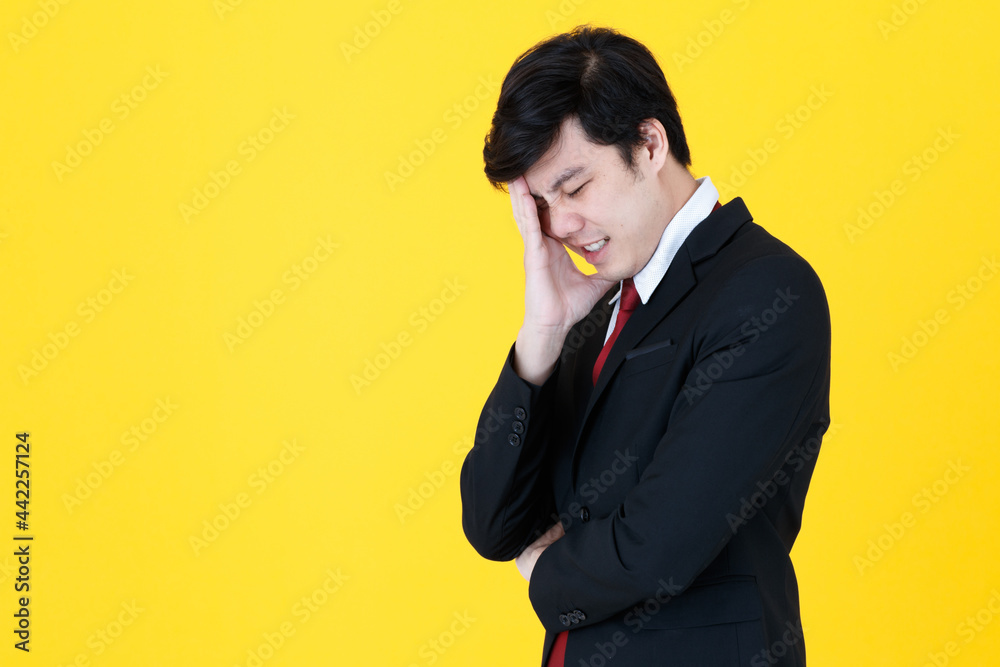 Horizontal portrait shot of serious Asian businessman in a black suit standing and touching this head with his hand to relieve stress isolated on yellow background. Concept of stress in the workplace Horizontal portrait shot of serious Asian businessman in a black suit standing and touching this head with his hand to relieve stress isolated on yellow background. Concept of stress in the workplace