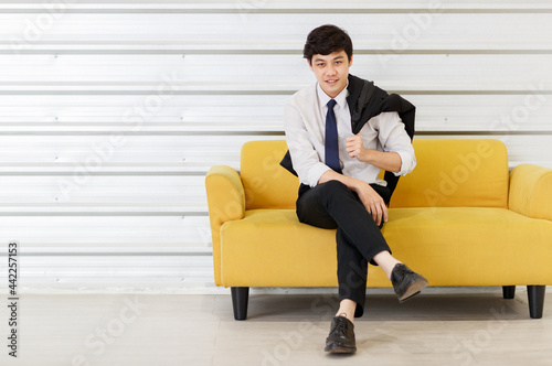 Horizontal portrait shot of young adult Asian handsome man in a white shirt with a suit on the shoulder sitting on the sofa and looking at the camera with hand to chin gesture posing in the studio